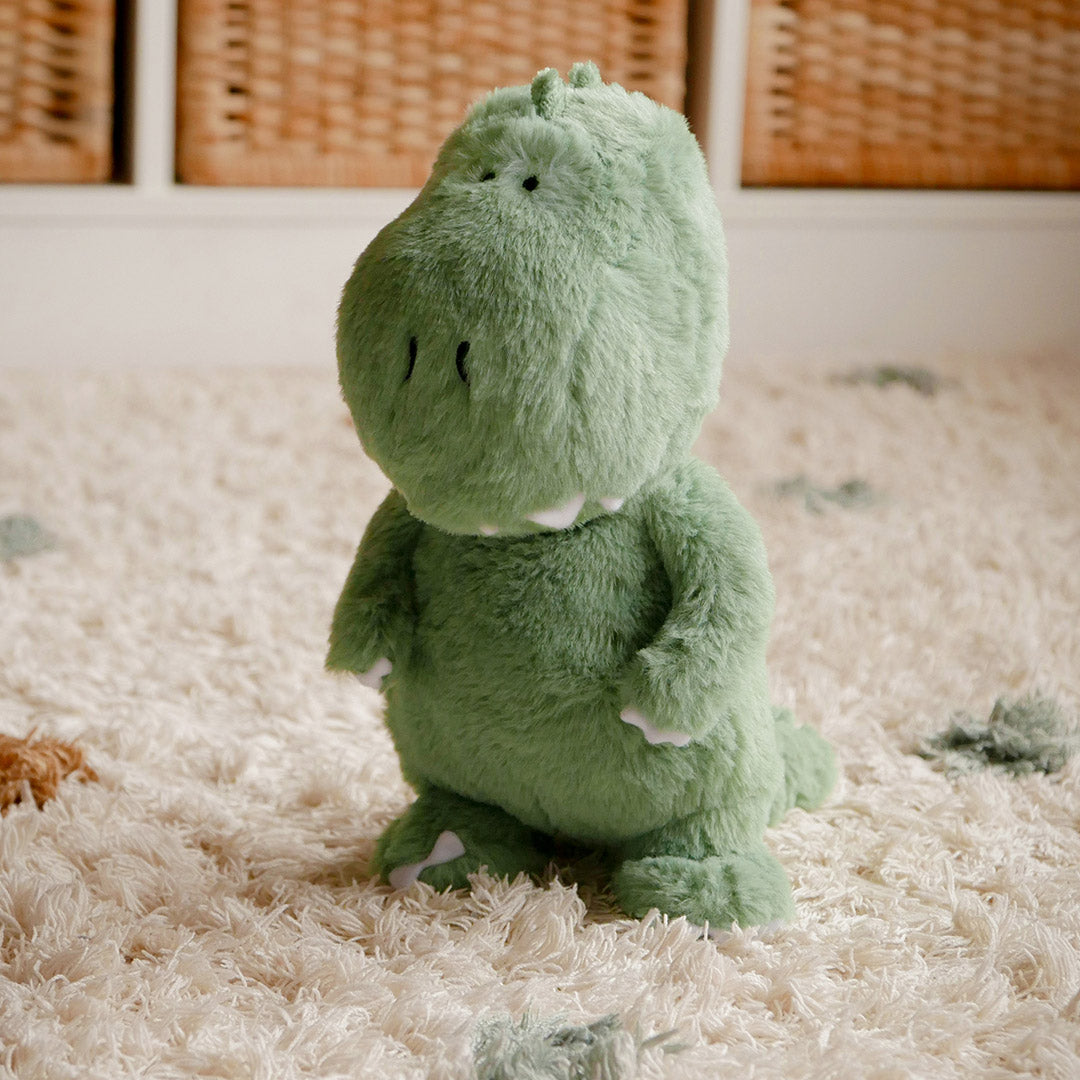 A green T-Rex dinosaur plush soft toy sitting on a soft, fluffy surface in a kids bedroom